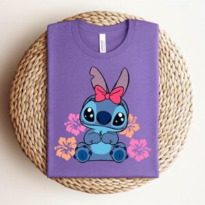 Puede incluir: Una camiseta morada con un personaje de dibujos animados con pelaje azul, ojos grandes y un lazo rosa. Flores de hibisco rosas y naranjas rodean al personaje. La camiseta est&aacute; doblada sobre una superficie tejida circular.