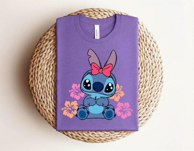 Puede incluir: Una camiseta morada con un personaje de dibujos animados con pelaje azul, ojos grandes y un lazo rosa. Flores de hibisco rosas y naranjas rodean al personaje. La camiseta est&aacute; doblada sobre una superficie tejida circular.