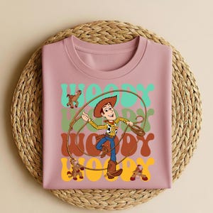 Peut inclure: Un t-shirt rose clair avec une illustration de Woody de Toy Story. Le design comprend Woody avec un lasso, le mot "WOODY" en police r&eacute;tro et des images de chevaux jouets. Le t-shirt est pr&eacute;sent&eacute; sur un tapis tiss&eacute;.
