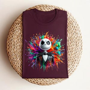 May include: A dark maroon t-shirt featuring a graphic print of a white skeletal character with a stitched mouth, wearing a black pinstripe suit and bow tie, centered against a vibrant, multi-colored paint splatter background with bright hues.