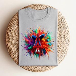 May include: A gray t-shirt featuring a colorful graphic of Spider-Man hanging upside down. The superhero is surrounded by a vibrant explosion of paint in shades of red, blue, green, and yellow. The shirt is folded neatly on a woven surface.
