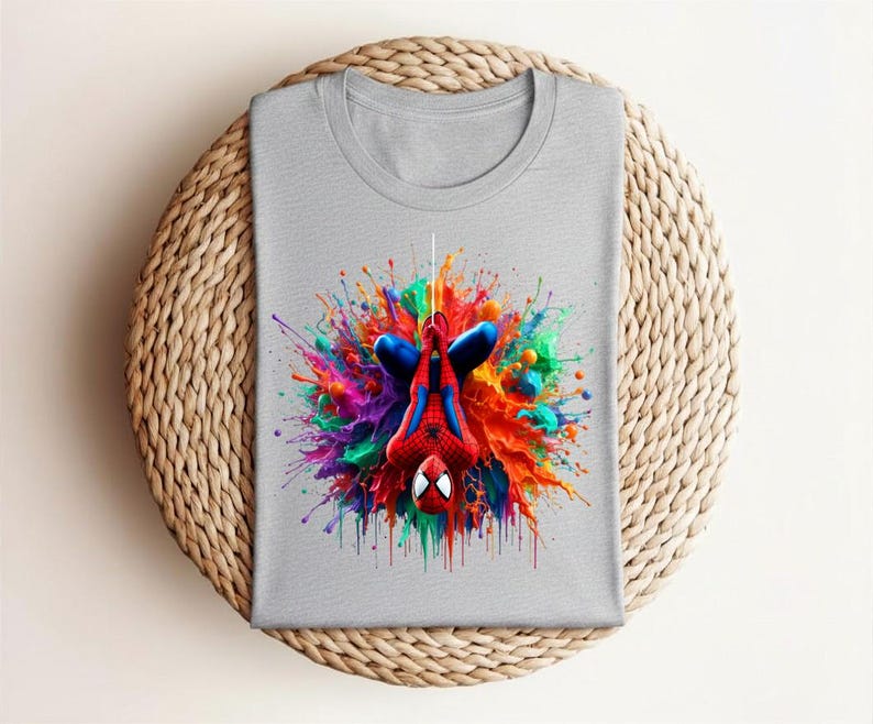 May include: A gray t-shirt featuring a colorful graphic of Spider-Man hanging upside down. The superhero is surrounded by a vibrant explosion of paint in shades of red, blue, green, and yellow. The shirt is folded neatly on a woven surface.