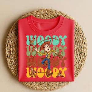 Peut inclure: T-shirt rouge &agrave; col rond avec une illustration de Woody de Toy Story. Woody tient un lasso, avec le mot "Woody" en police r&eacute;tro. Le design comprend un cheval et deux autres figurines.