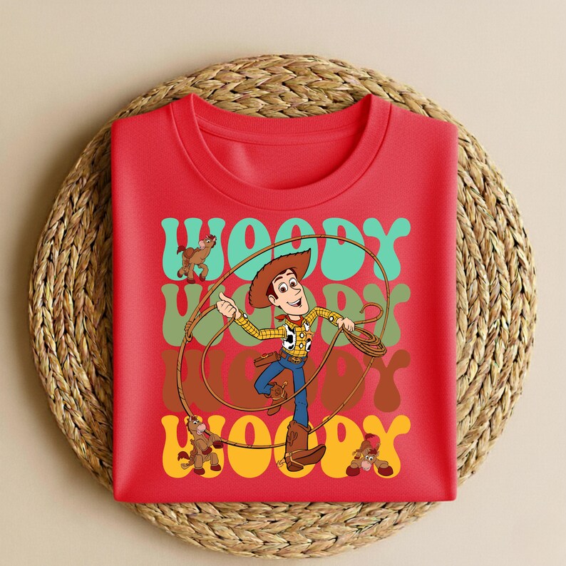 Peut inclure: T-shirt rouge &agrave; col rond avec une illustration de Woody de Toy Story. Woody tient un lasso, avec le mot "Woody" en police r&eacute;tro. Le design comprend un cheval et deux autres figurines.
