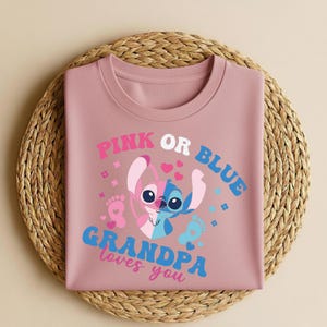 Pu&ograve; includere: Una maglietta color rosa antico con la scritta "PINK OR BLUE GRANDPA loves you". La grafica presenta un personaggio dei cartoni animati bicolore, rosa e blu, con cuori e impronte. La maglietta &egrave; piegata su un sottobicchiere circolare intrecciato.