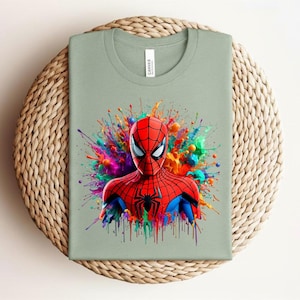 Op de afbeelding: Saliegroen t-shirt met een levendige illustratie van Spider-Man. De superheld is afgebeeld in een rood en blauw pak, tegen een kleurrijke verfspat. Het shirt is op een geweven oppervlak gevouwen.