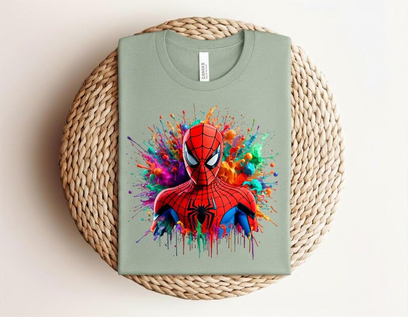 Op de afbeelding: Saliegroen t-shirt met een levendige illustratie van Spider-Man. De superheld is afgebeeld in een rood en blauw pak, tegen een kleurrijke verfspat. Het shirt is op een geweven oppervlak gevouwen.