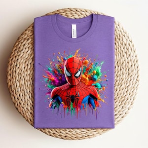 Op de afbeelding: Een paars t-shirt met een kleurrijke afbeelding van Spider-Man. De superheld is afgebeeld in een rood en zwart pak, tegen een levendige spetter van verf in verschillende kleuren. Het shirt is op een geweven oppervlak gevouwen.