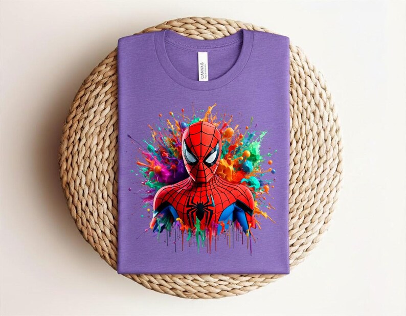 Op de afbeelding: Een paars t-shirt met een kleurrijke afbeelding van Spider-Man. De superheld is afgebeeld in een rood en zwart pak, tegen een levendige spetter van verf in verschillende kleuren. Het shirt is op een geweven oppervlak gevouwen.