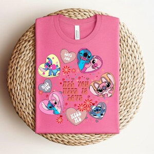 Peut inclure: Un t-shirt rose avec des personnages de dessins anim&eacute;s et des motifs en forme de c&oelig;ur. Le t-shirt porte l'inscription "ARE YOU HAPPY IS LOVE" et "LOVE YOU", "MISS YOU" et "KISS ME" sur les c&oelig;urs. Le t-shirt est pli&eacute; sur une surface tiss&eacute;e.