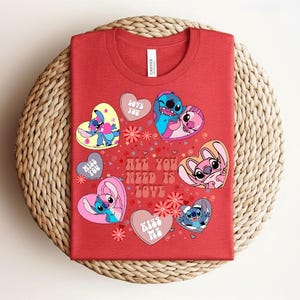 Peut inclure: T-shirt rouge orn&eacute; de graphismes en forme de c&oelig;ur avec des personnages de dessins anim&eacute;s et le texte "ALL YOU NEED IS LOVE". Le design comprend des personnages dans des poses amoureuses, avec des textes comme "LOVE YOU" et "KISS ME".