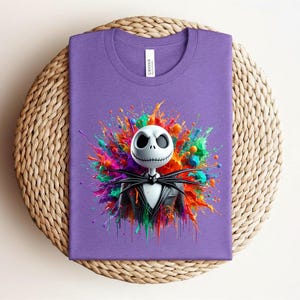 May include: A purple t-shirt featuring a graphic of a white skull character with a stitched mouth wearing a black pinstripe suit and bow tie, centered against a vibrant, multi-colored paint splatter design with splashes of red, blue, green, and orange.