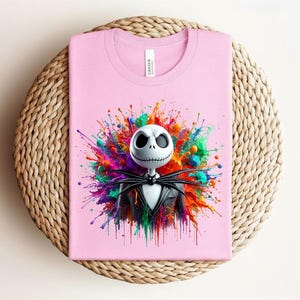 May include: A light pink t-shirt featuring a graphic of a white skull character with large black eyes and a stitched mouth, wearing a black pinstripe suit and bow tie, set against a vibrant, multi-colored paint splatter background.