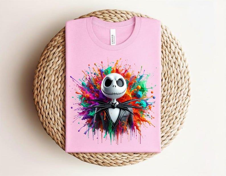 May include: A light pink t-shirt featuring a graphic of a white skull character with large black eyes and a stitched mouth, wearing a black pinstripe suit and bow tie, set against a vibrant, multi-colored paint splatter background.