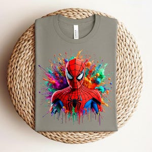 Op de afbeelding: Een grijs t-shirt met een kleurrijke afbeelding van Spider-Man. De superheld wordt afgebeeld in een rood en blauw pak, tegen een levendige spetter van verf in verschillende kleuren. Het shirt wordt getoond op een geweven, cirkelvormig oppervlak.