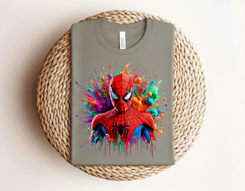 Op de afbeelding: Een grijs t-shirt met een kleurrijke afbeelding van Spider-Man. De superheld wordt afgebeeld in een rood en blauw pak, tegen een levendige spetter van verf in verschillende kleuren. Het shirt wordt getoond op een geweven, cirkelvormig oppervlak.