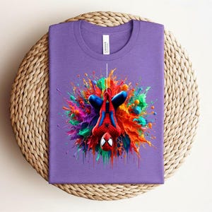 May include: A purple t-shirt featuring a colorful graphic of Spider-Man in a dynamic pose. The design showcases the superhero with vibrant splashes of red, blue, orange, and green, creating a bold and eye-catching visual. The shirt is folded neatly on a woven surface.