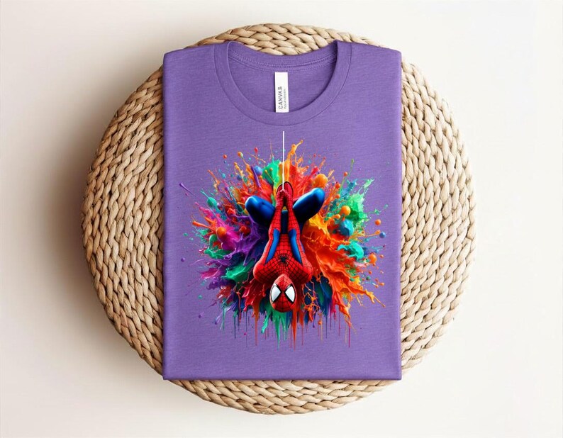 May include: A purple t-shirt featuring a colorful graphic of Spider-Man in a dynamic pose. The design showcases the superhero with vibrant splashes of red, blue, orange, and green, creating a bold and eye-catching visual. The shirt is folded neatly on a woven surface.