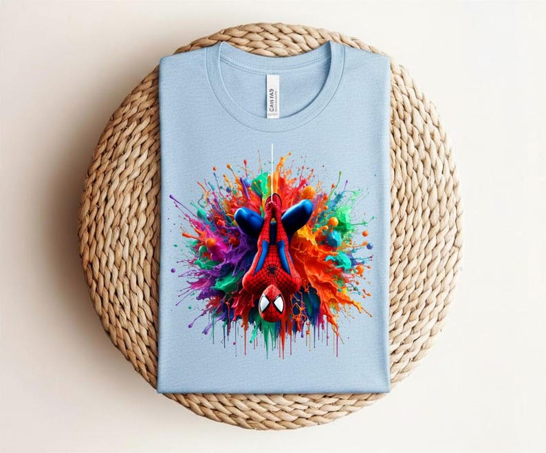 May include: A light blue t-shirt featuring a colorful graphic of Spider-Man hanging upside down. The superhero is depicted in his classic red and blue suit, surrounded by a vibrant explosion of paint in various colors. The shirt is made of a soft material.