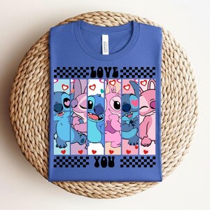 May include: Blue t-shirt featuring a graphic of cartoon characters in shades of blue and pink, with the words "LOVE YOU" in black. The design includes hearts and a checkered pattern.