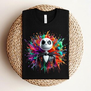 May include: A black t-shirt featuring a graphic of a white skeletal character with a stitched mouth and large eye sockets, wearing a black pinstripe suit and bow tie, set against a vibrant, multi-colored paint splatter background.