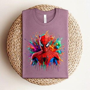 Op de afbeelding: Een mauvekleurig T-shirt met een levendige afbeelding van Spider-Man. De superheld is afgebeeld in zijn rood-blauwe pak, tegen een kleurrijke verfspat. Het T-shirt is opgevouwen op een ronde, geweven mat.