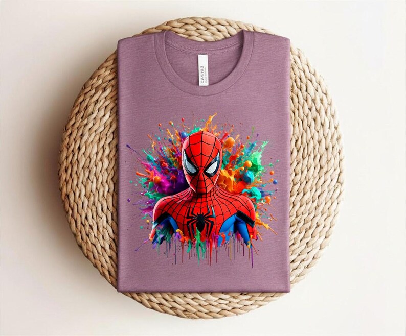 Op de afbeelding: Een mauvekleurig T-shirt met een levendige afbeelding van Spider-Man. De superheld is afgebeeld in zijn rood-blauwe pak, tegen een kleurrijke verfspat. Het T-shirt is opgevouwen op een ronde, geweven mat.