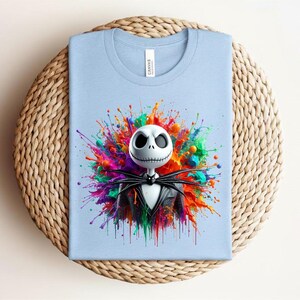 May include: A light blue t-shirt featuring a graphic of a white skull character with large black eyes and a stitched mouth, wearing a black pinstripe suit and bow tie, centered against a vibrant, multi-colored paint splatter background.