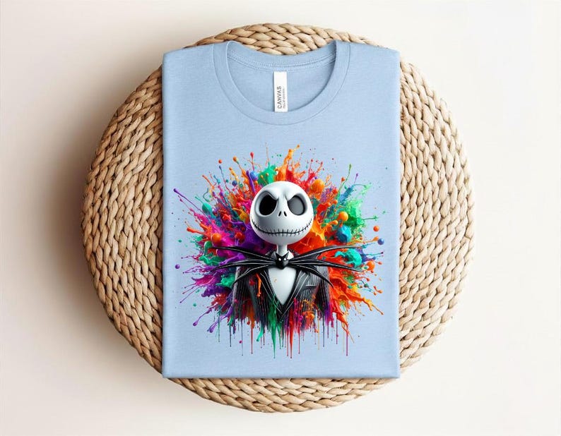 May include: A light blue t-shirt featuring a graphic of a white skull character with large black eyes and a stitched mouth, wearing a black pinstripe suit and bow tie, centered against a vibrant, multi-colored paint splatter background.