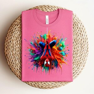 May include: A pink t-shirt featuring a colorful graphic of Spider-Man hanging upside down. The superhero is surrounded by a vibrant explosion of paint in shades of blue, red, orange, yellow, green, and purple. The shirt is folded on a woven surface.