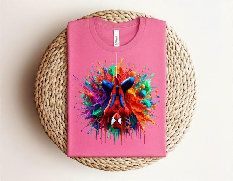 May include: A pink t-shirt featuring a colorful graphic of Spider-Man hanging upside down. The superhero is surrounded by a vibrant explosion of paint in shades of blue, red, orange, yellow, green, and purple. The shirt is folded on a woven surface.