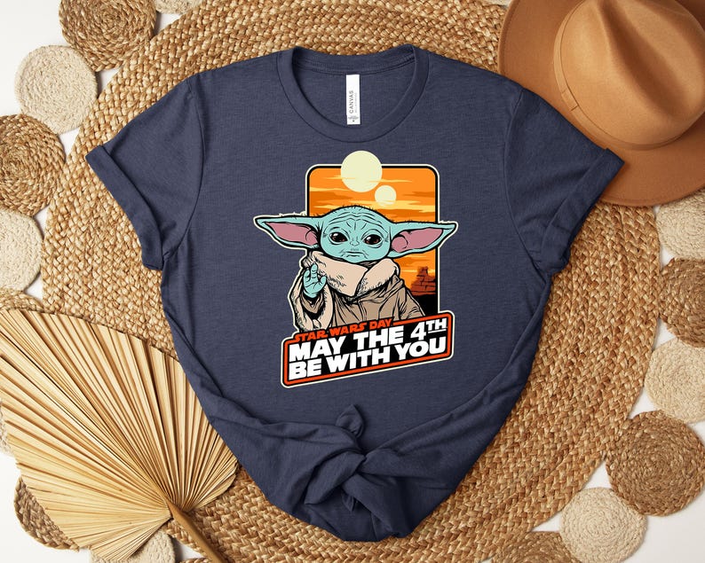 Może przedstawiać: Granatowa koszulka z grafiką Grogu z Gwiezdnych Wojen i napisem "STAR WARS DAY MAY THE 4TH BE WITH YOU". Projekt zawiera pomarańczowy zach&oacute;d słońca i dwa słońca. Koszulka ma okrągły dekolt.