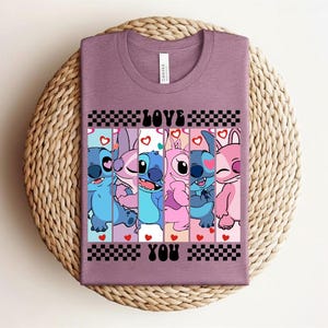 May include: Mauve t-shirt featuring a graphic of cartoon characters with the words "LOVE YOU" in black. The design includes blue, pink, and white characters with hearts and checkered patterns. The shirt is displayed on a woven, circular surface.