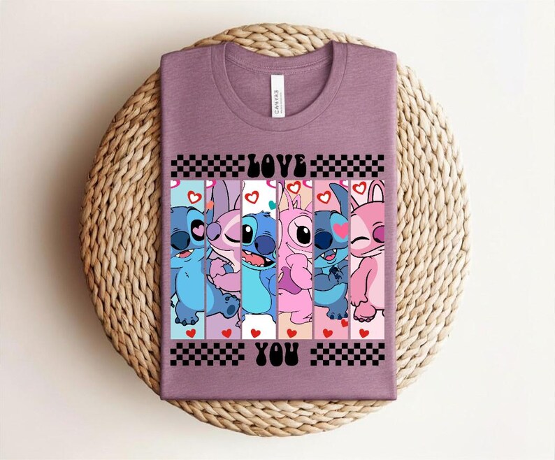 May include: Mauve t-shirt featuring a graphic of cartoon characters with the words "LOVE YOU" in black. The design includes blue, pink, and white characters with hearts and checkered patterns. The shirt is displayed on a woven, circular surface.