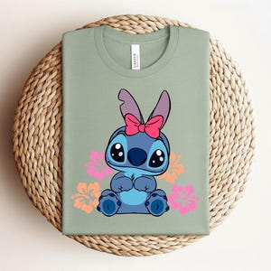 Puede incluir: Camiseta verde salvia con un personaje de dibujos animados azul, con un lazo rosa y rodeado de flores rosas y naranjas. La camiseta est&aacute; doblada sobre una superficie circular tejida. La camiseta es de algod&oacute;n.