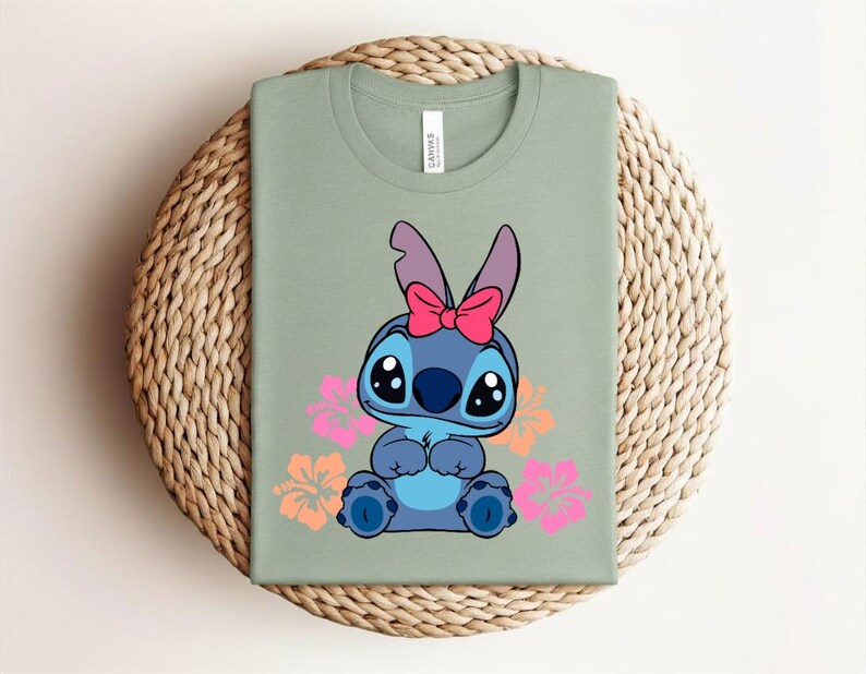 Puede incluir: Camiseta verde salvia con un personaje de dibujos animados azul, con un lazo rosa y rodeado de flores rosas y naranjas. La camiseta est&aacute; doblada sobre una superficie circular tejida. La camiseta es de algod&oacute;n.