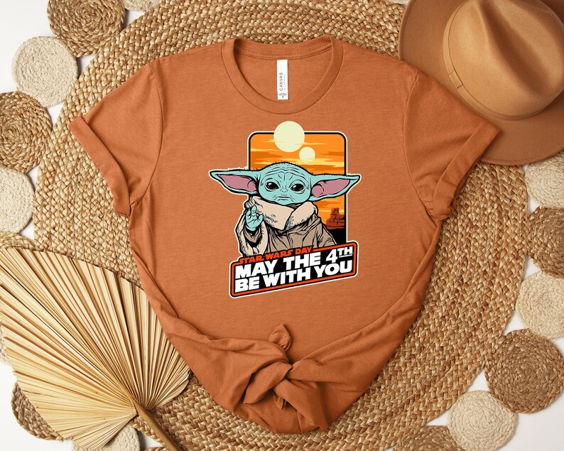 Może przedstawiać: Rdzawa koszulka z grafiką Grogu z Gwiezdnych Wojen. Obraz zawiera tekst "Star Wars Day May the 4th Be With You". Projekt przedstawia Grogu z dużymi uszami i beżowym płaszczem na tle pomarańczowego zachodu słońca.