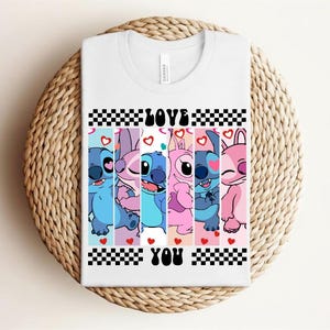 May include: White t-shirt featuring a colorful graphic of cartoon characters with the words "LOVE YOU" in black. The design includes blue, pink, and purple characters with hearts and a checkered pattern.