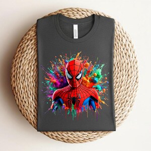 Op de afbeelding: Donkergrijs t-shirt met een levendige illustratie van Spider-Man. De superheld is afgebeeld in zijn rood-blauwe pak, tegen een kleurrijke verfspat. Het t-shirt is op een geweven oppervlak gevouwen.
