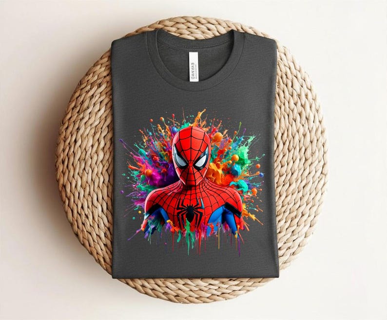 Op de afbeelding: Donkergrijs t-shirt met een levendige illustratie van Spider-Man. De superheld is afgebeeld in zijn rood-blauwe pak, tegen een kleurrijke verfspat. Het t-shirt is op een geweven oppervlak gevouwen.