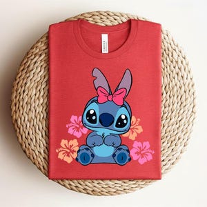 Puede incluir: Camiseta roja con un personaje de dibujos animados azul, con un lazo rosa y rodeado de flores de hibisco rosas y naranjas. La camiseta est&aacute; doblada sobre una superficie tejida.