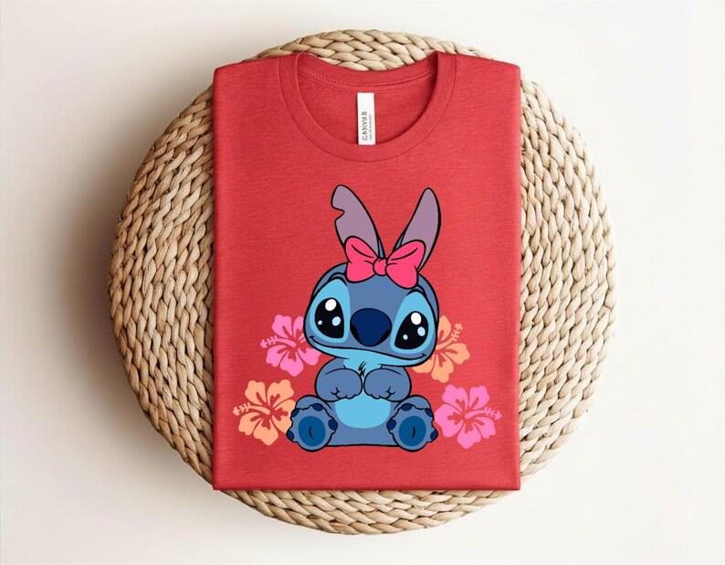Puede incluir: Camiseta roja con un personaje de dibujos animados azul, con un lazo rosa y rodeado de flores de hibisco rosas y naranjas. La camiseta est&aacute; doblada sobre una superficie tejida.