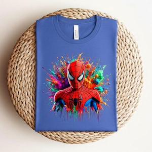 Op de afbeelding: Een blauw t-shirt met een kleurrijke afbeelding van Spider-Man. Het gezicht en bovenlichaam van de superheld zijn afgebeeld in rood en zwart, tegen een levendige splash van regenboogkleuren. Het shirt ligt plat op een geweven oppervlak.