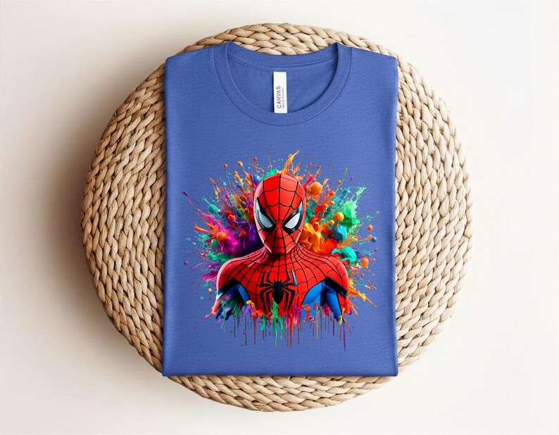 Op de afbeelding: Een blauw t-shirt met een kleurrijke afbeelding van Spider-Man. Het gezicht en bovenlichaam van de superheld zijn afgebeeld in rood en zwart, tegen een levendige splash van regenboogkleuren. Het shirt ligt plat op een geweven oppervlak.