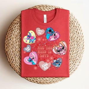 Peut inclure: T-shirt rouge avec des personnages de dessins anim&eacute;s en forme de c&oelig;ur, avec les phrases "Love You" et "Kiss Me". Le t-shirt porte &eacute;galement l'inscription "All You Need Is Love". Le t-shirt est pr&eacute;sent&eacute; sur une surface tiss&eacute;e.