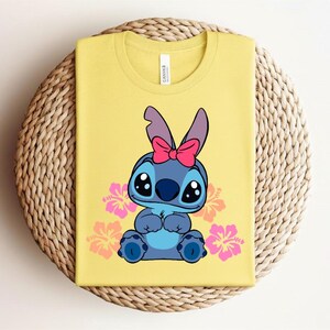 Puede incluir: Camiseta amarilla con un personaje de dibujos animados con pelaje azul, ojos grandes y un lazo rosa. Flores de hibisco rosas y naranjas rodean al personaje. La camiseta est&aacute; doblada sobre una superficie circular tejida.