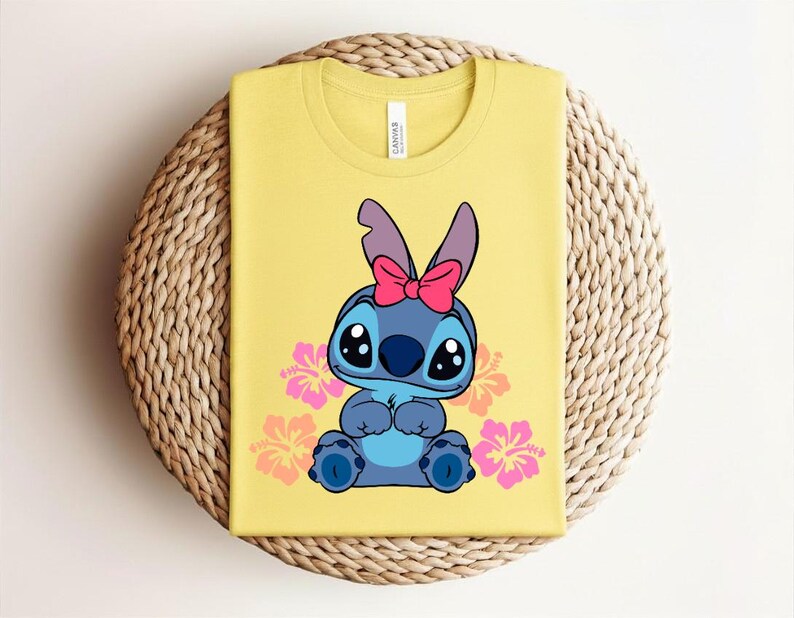 Puede incluir: Camiseta amarilla con un personaje de dibujos animados con pelaje azul, ojos grandes y un lazo rosa. Flores de hibisco rosas y naranjas rodean al personaje. La camiseta est&aacute; doblada sobre una superficie circular tejida.