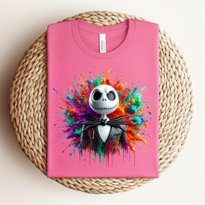 May include: A pink t-shirt featuring a graphic of a white skull character with a black pinstripe suit and bow tie, centered against a vibrant, multi-colored paint splatter design. The shirt has a crew neckline and a white brand label at the collar.