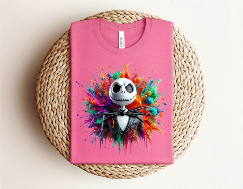 May include: A pink t-shirt featuring a graphic of a white skull character with a black pinstripe suit and bow tie, centered against a vibrant, multi-colored paint splatter design. The shirt has a crew neckline and a white brand label at the collar.