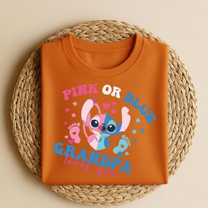 Pu&ograve; includere: Una maglietta arancione con la scritta "PINK OR BLUE GRANDPA loves you" e un personaggio dei cartoni animati rosa e blu. La maglietta &egrave; piegata su un sottobicchiere circolare intrecciato.